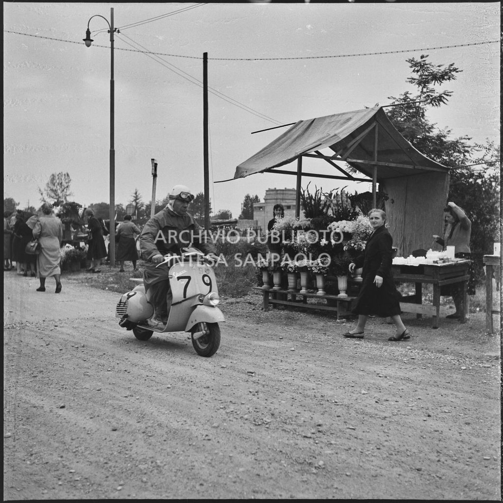 Un concorrente(numero di gara 79) passa accanto ad un fiorista durante la competizione sportiva 1000 Km Vespistica del 1954<br>216540