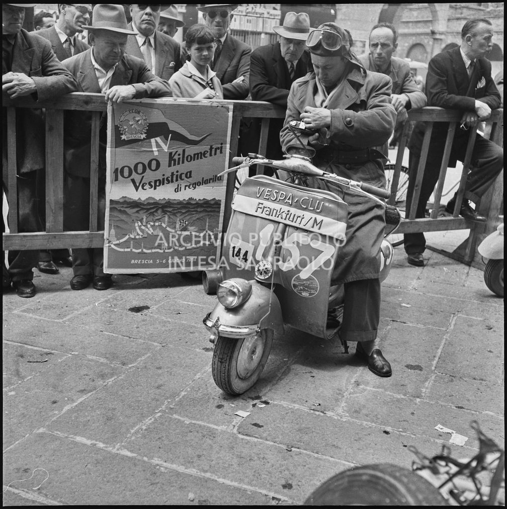 Un concorrente del Vespa Club di Francoforte (numero di gara 157), con in mano una macchina fotografica, in piazza della Vittoria a Brescia in occasione della competizione sportiva 1000 Km Vespistica del 1954<br>216590