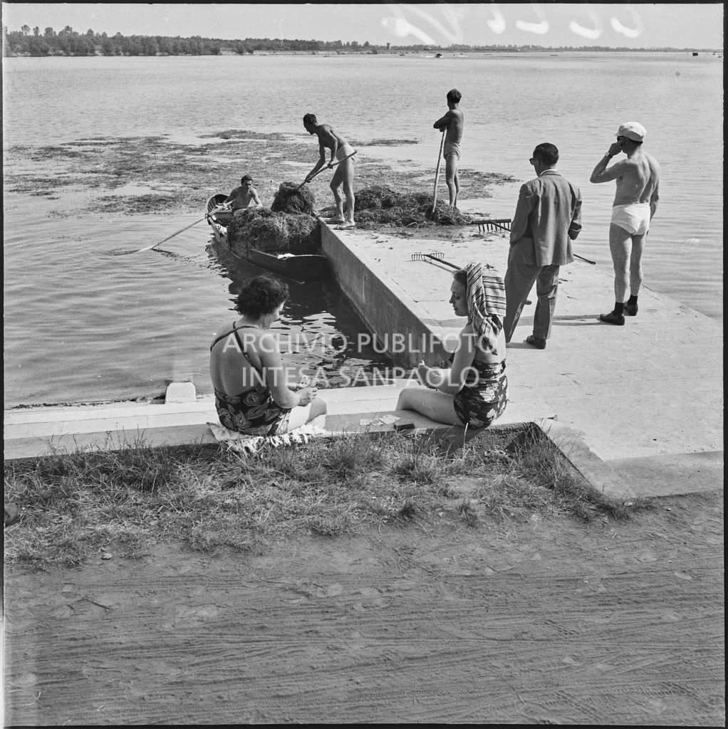 Alcuni uomini raccolgono delle erbacce dalle acque dell'Idroscalo di Milano mentre due donne giocano a carte sulla riva<br>222196