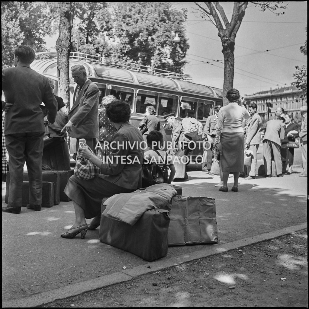 Passeggeri seduti sulle valige sul ciglio di una strada di Milano in attesa della partenza del pullman per le vacanze estive per la Presolana<br>222874