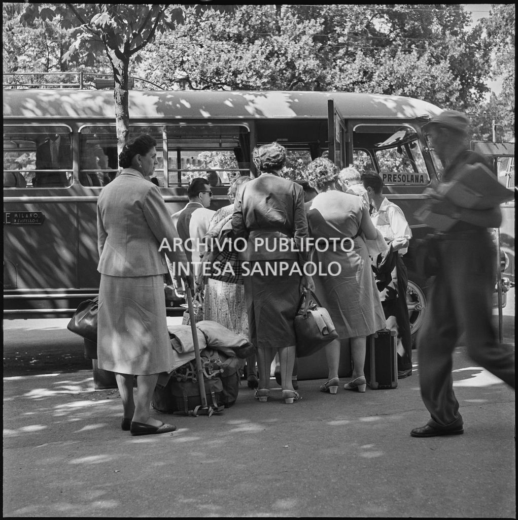 Passeggeri seduti sulle valige sul ciglio di una strada di Milano in attesa della partenza del pullman per le vacanze estive per la Presolana<br>222874 bis