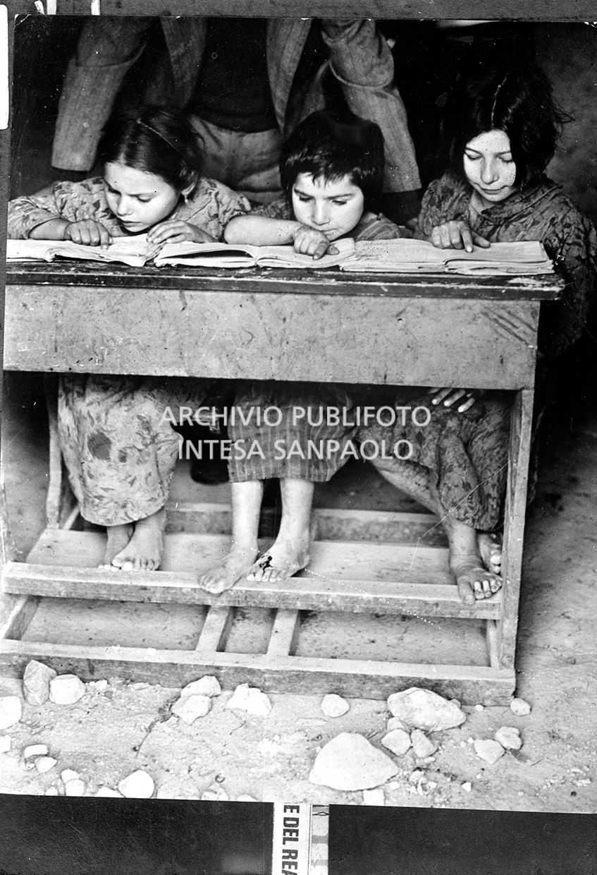 Africo, bambine scalze sedute a un banco di scuola intente alla lettura; alle loro spalle il maestro segue l'esercitazione