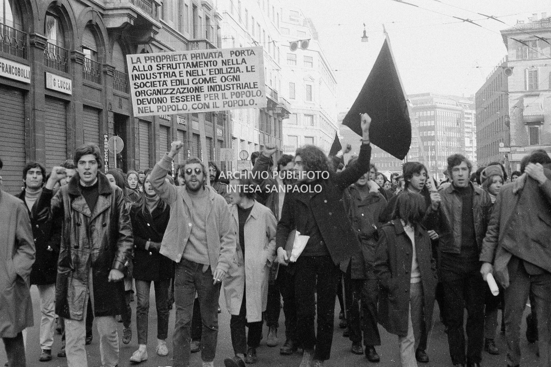 Manifestazione contro il caro casa nel giorno dello sciopero generale; un cartello recita: "La proprietà privata porta allo sfruttamento nell'industria e nell'edilizia. Le società edili come ogni organizzazione industriale devono essere per il popolo, nel popolo, con il popolo."