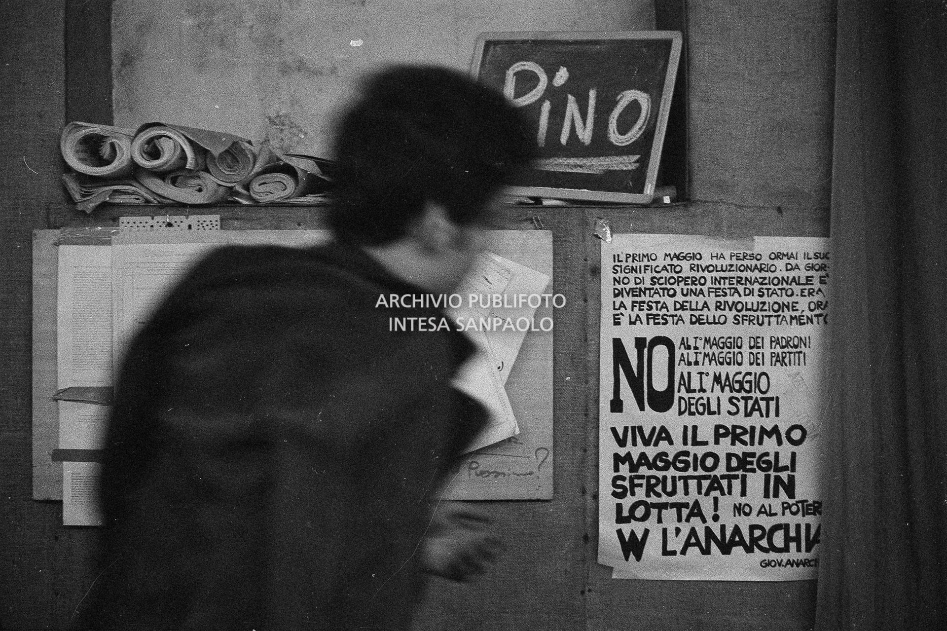 Un uomo di spalle al circolo anarchico Ponte della Ghisolfa; dietro di lui su una lavagnetta la scritta "Pino"