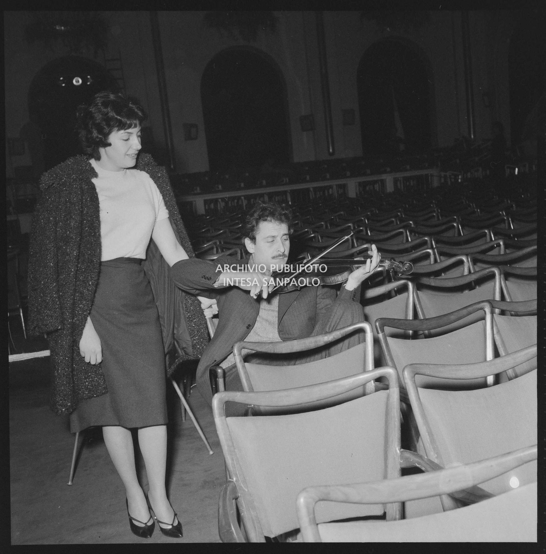 Domenico Modugno, seduto nella platea di un teatro, suona il violino, in compagnia della moglie Franca Gandolfi