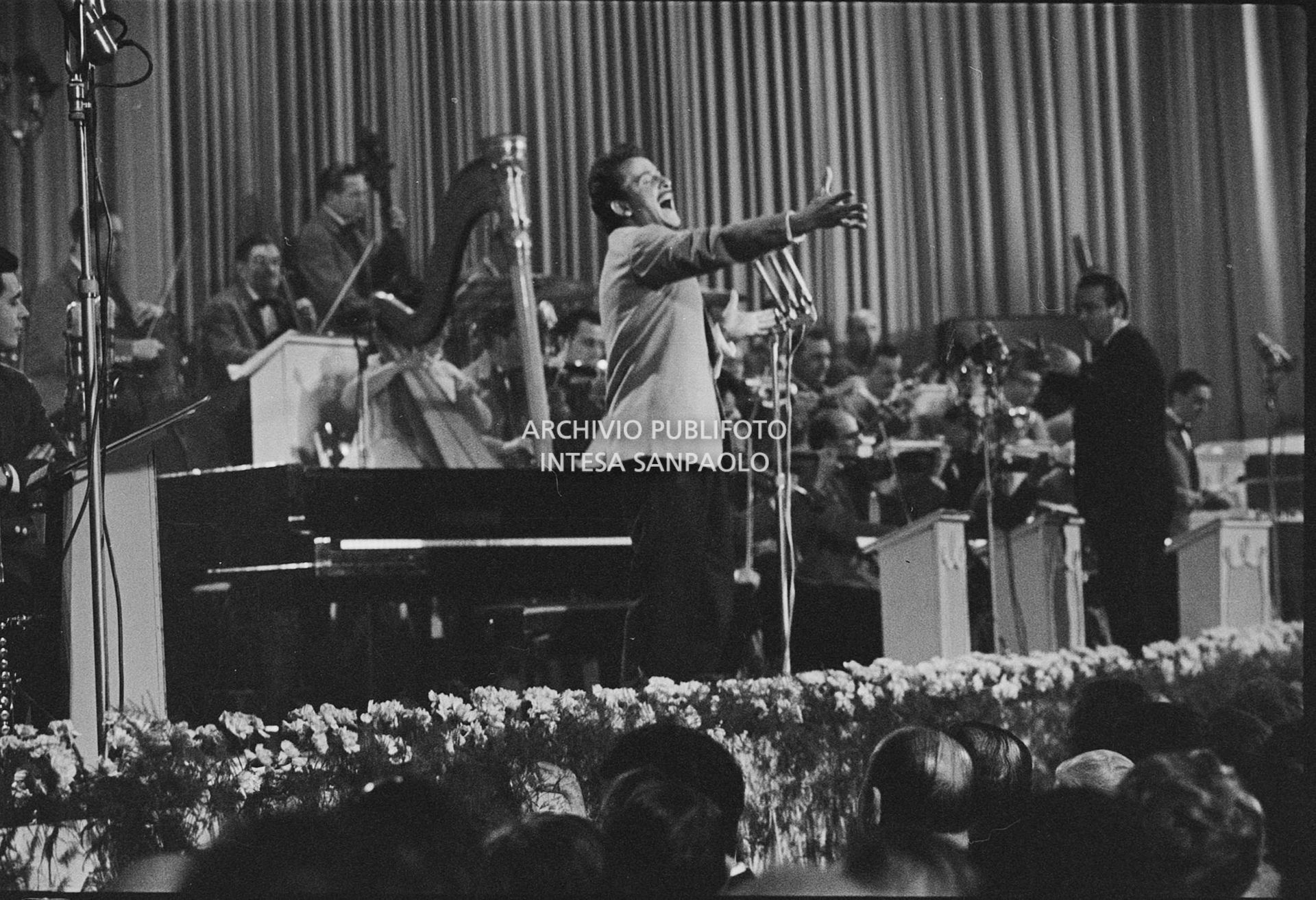 Domenico Modugno, si esibisce sul palco del Salone delle feste del Casinò al IX Festival di Sanremo, che vincerà, insieme a Johnny Dorelli, con la canzone "Piove"