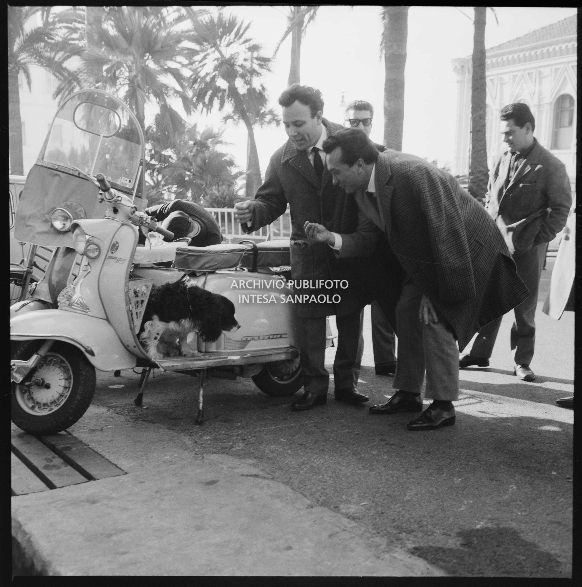 Claudio Villa e Ernesto Bonino si rivolgono a un cagnolino a bordo di una Lambretta parcheggiata davanti al Casinò municipale nei giorni del XII Festival di Sanremo