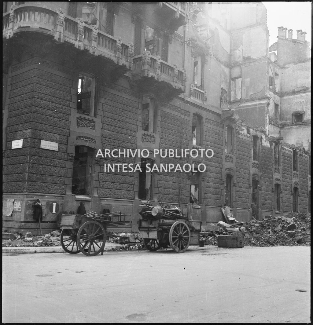 Esterno dell'edificio in viale Piave 15 all'angolo con via Lamberto De Bernardi, a Milano, sventrato dai bombardamenti; per strada macerie e due carretti a trazione umana. Sul muro il segnale "I" che indica l'attacco per l'idrante.