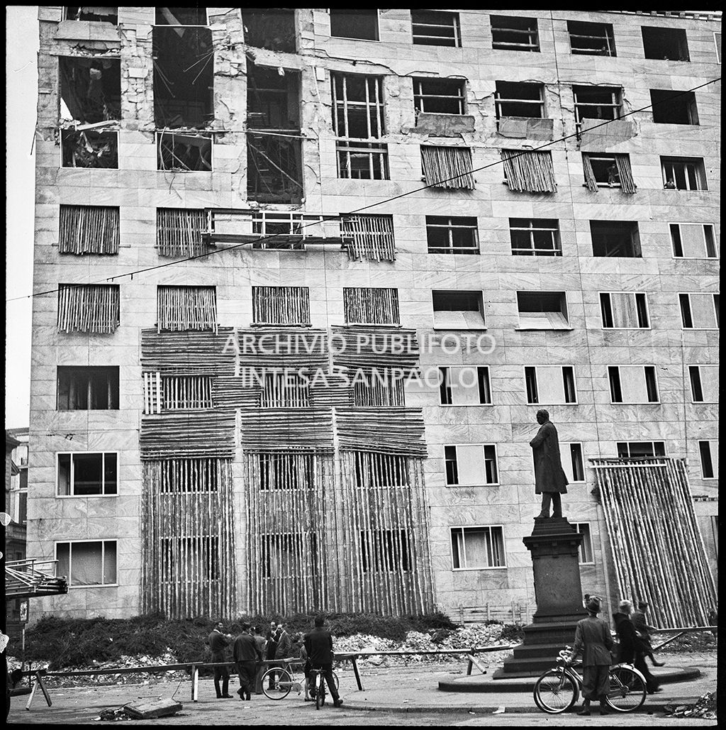 Passanti e militari davanti a Palazzo Montecatini, in via della Moscova a Milano, gravemente danneggiato dai bombardamenti; alcune opere di messa in sicurezza sono già state apprestate, i piani superiori sono sventrati e un pilastro è pericolante. In basso a destra il monumento a Agostino Bertani