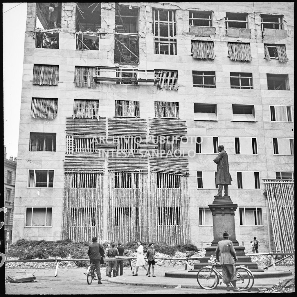 Passanti e militari davanti a Palazzo Montecatini, in via della Moscova a Milano, gravemente danneggiato dai bombardamenti; alcune opere di messa in sicurezza sono già state apprestate, i piani superiori sono sventrati e un pilastro è pericolante. In basso a destra il monumento a Agostino Bertani