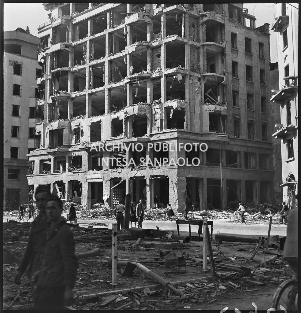 Edificio in largo Augusto all'angolo con via Marziale, a Milano, sventrato dai bombardamenti; in primo piano due militari, sullo sfondo 
				passanti tra le macerie