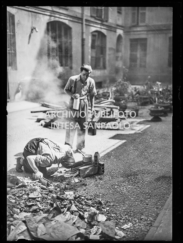 Due uomini cucinano il pranzo su un focolare di fortuna, allestito per strada a Milano, tra le macerie degli edifici bombardati
