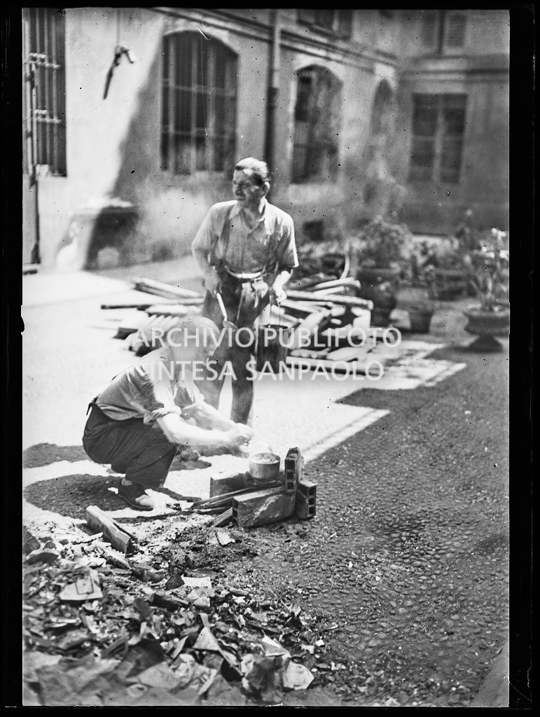 Due uomini cucinano il pranzo su un focolare di fortuna, allestito per strada a Milano, tra le macerie degli edifici bombardati