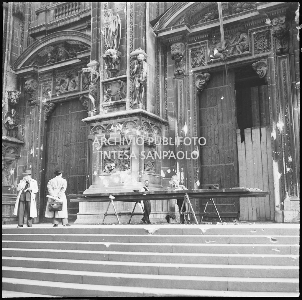 Pannello a protezione del portale del Duomo di Milano, danneggiato durante i bombardamenti, in riparazione sul sagrato del Duomo. Sulla sinistra il portale minore anch'esso protetto da un pannello di legno