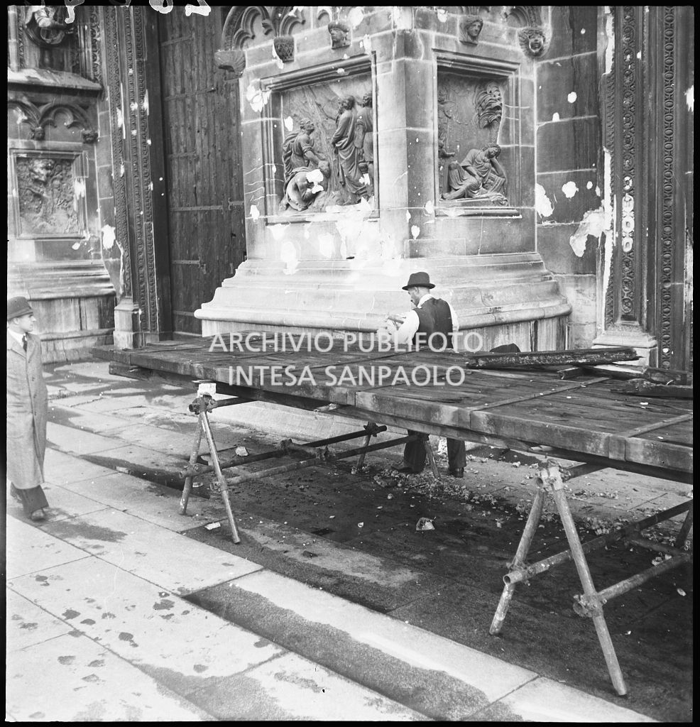 Pannello a protezione del portale del Duomo di Milano, danneggiato durante i bombardamenti, in riparazione sul sagrato del Duomo. Sulla sinistra il portale minore anch'esso protetto da un pannello di legno