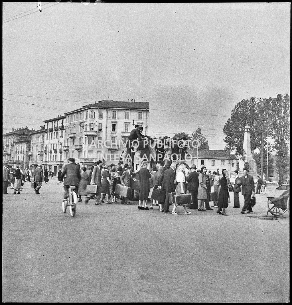 Una folla di persone, molte con valigia alla mano, si accalca per salire su un furgone in uscita da Milano dopo che la città è stata bombardata. Altri si avviano in bicicletta