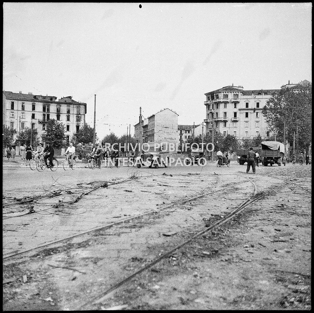 Scorcio di una piazza di Milano, con automobili e numerose persone in bicicletta, nei giorni dei bombardamenti della città