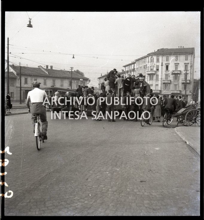 Una folla di persone, molte con valigia alla mano, si accalca per salire su un furgone in uscita da Milano dopo che la città è stata bombardata. Altri si avviano in bicicletta