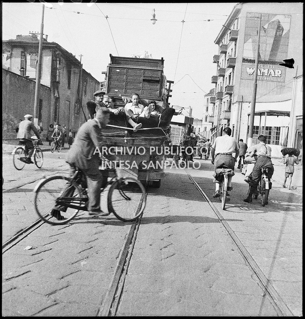 Esodo dalla città, dopo i bombardamenti, in via Carlo Farini, a Milano, con carri, trainati da cavalli, carichi di masserizie o in bicicletta. Sul muro a destra una grande pubblicità murale dei biscotti Wamar