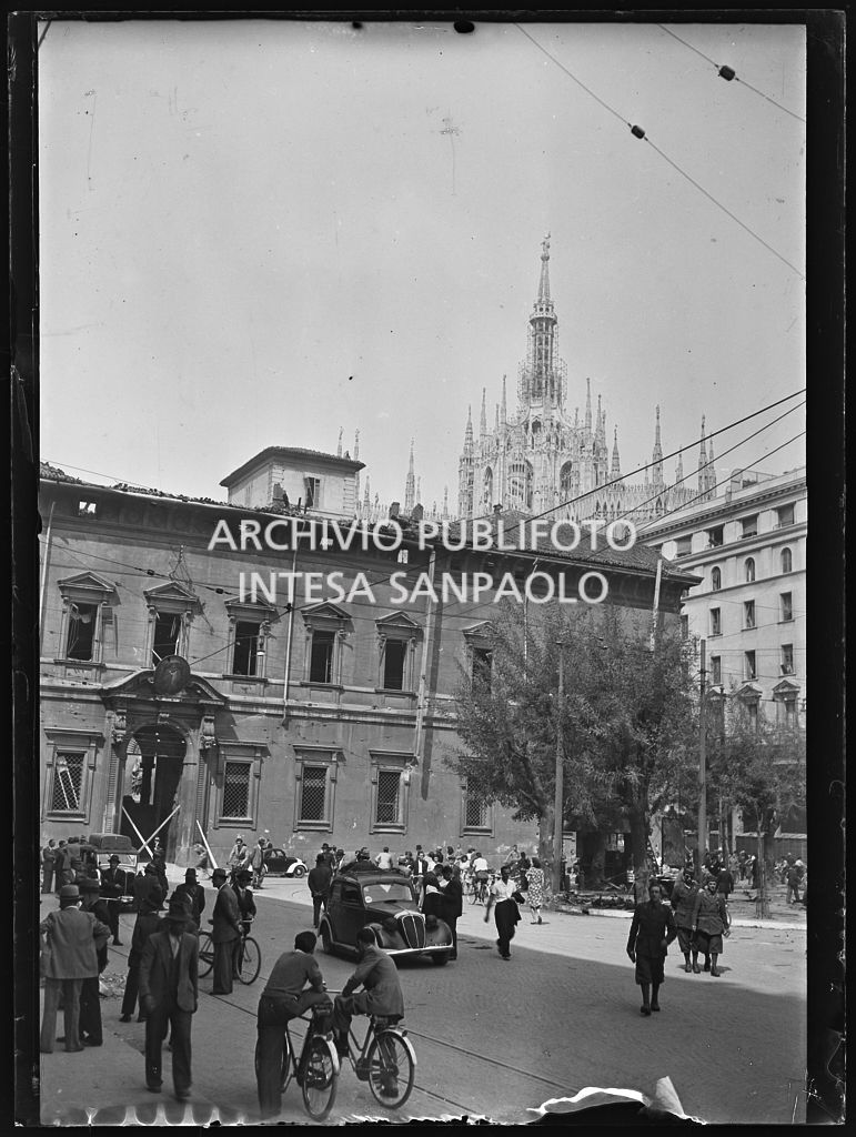 Passanti a piedi, in bicicletta e in automobile in piazza Fontana, all'indomani dei bombardamenti che hanno danneggiato il Palazzo Arcivescovile. Sullo sfondo le guglie del Duomo
