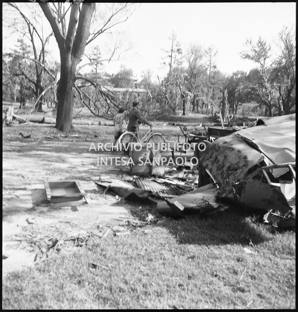Due passanti, uno con bicicletta, passeggiano ai Giardini Pubblici dopo i bombardamenti che hanno divelto numerosi alberi