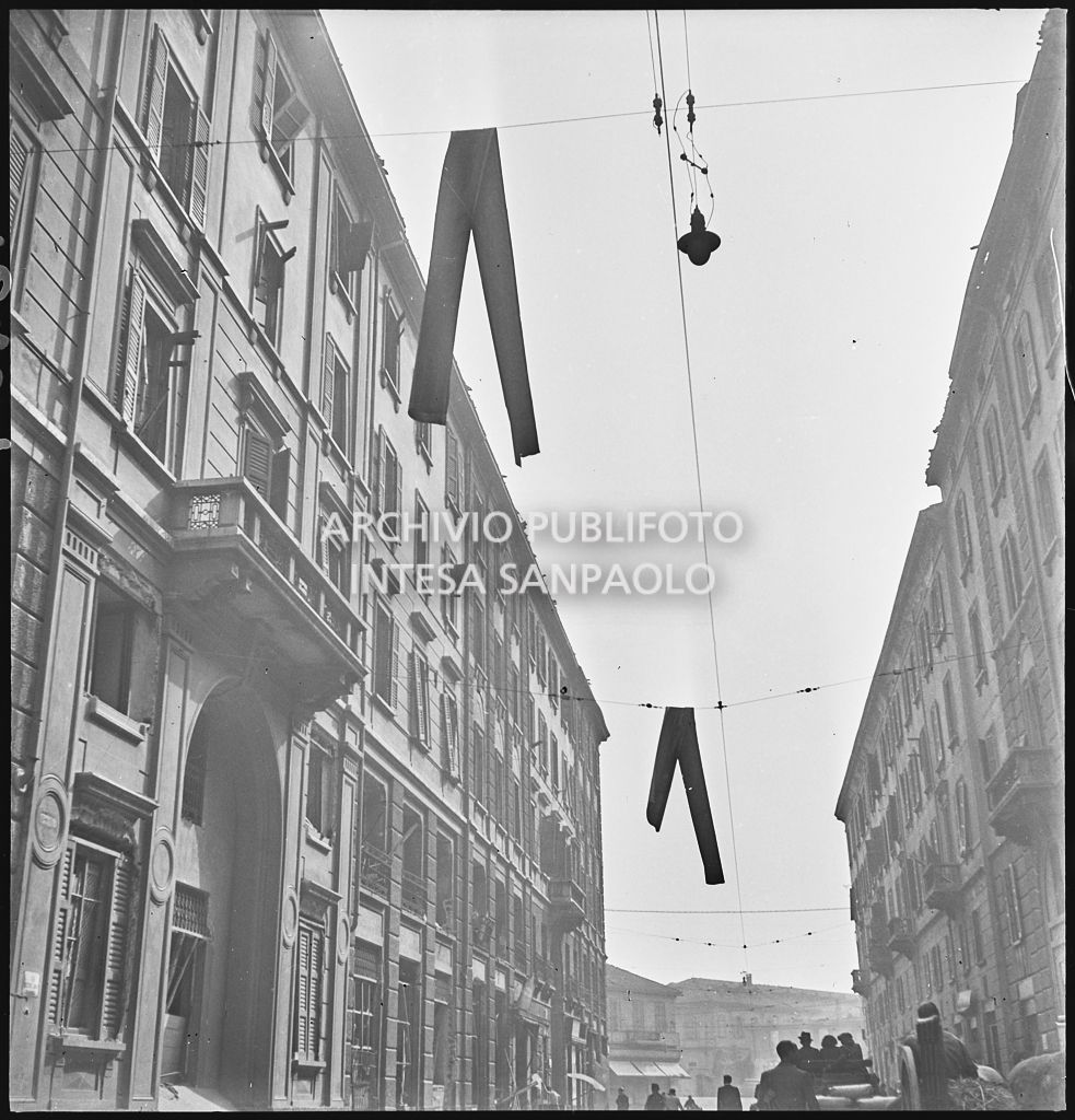 Passanti in una via di Milano all'indomani dei bombardamenti che hanno danneggiato gli edifici facendo cadere, sui fili della illuminazione cittadina, due grondaie