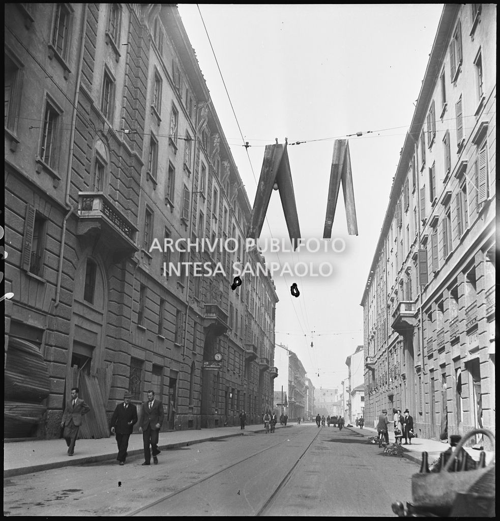 Passanti in una via di Milano all'indomani dei bombardamenti che hanno danneggiato gli edifici facendo cadere, sui fili della illuminazione cittadina, due grondaie