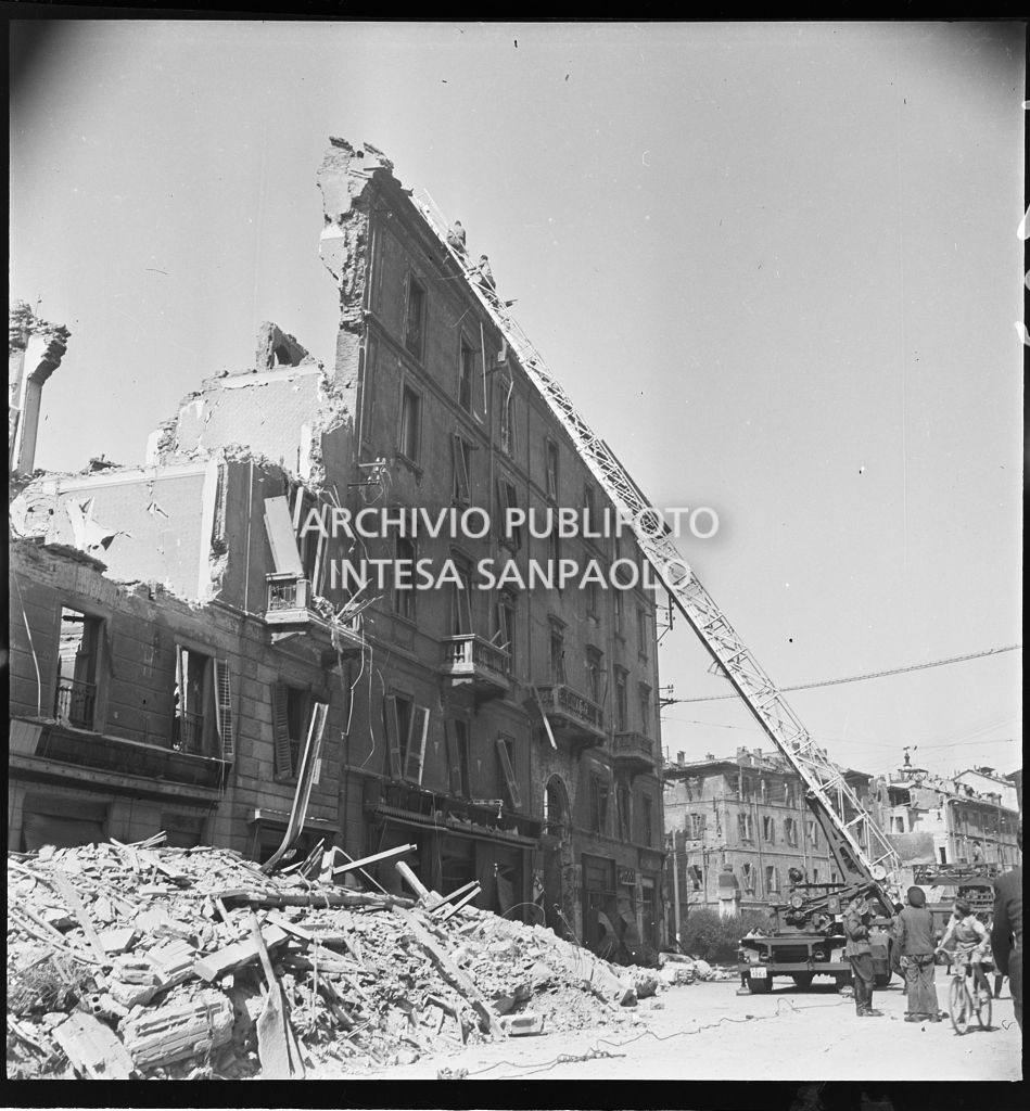 Due uomini verificano, con l'ausilio di un'autoscala, la stabilità di un edificio bombardato, in corso Genova a Milano, di cui ha resistito solo la facciata. A terra, addossati ai lati della strada, mucchi di macerie
