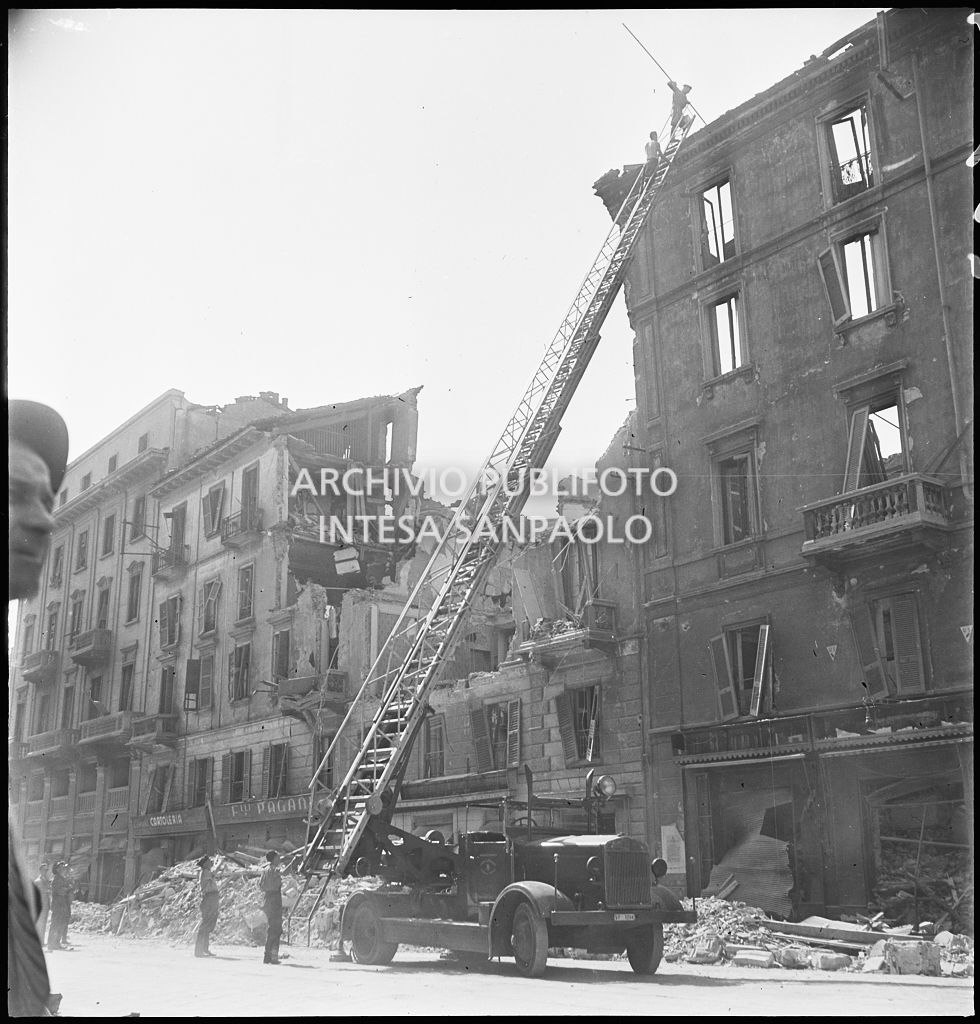 Due uomini verificano, con l'ausilio di un'autoscala, la stabilità di un edificio bombardato, in corso Genova a Milano, di cui ha resistito solo la facciata. A terra, addossati ai lati della strada, mucchi di macerie
