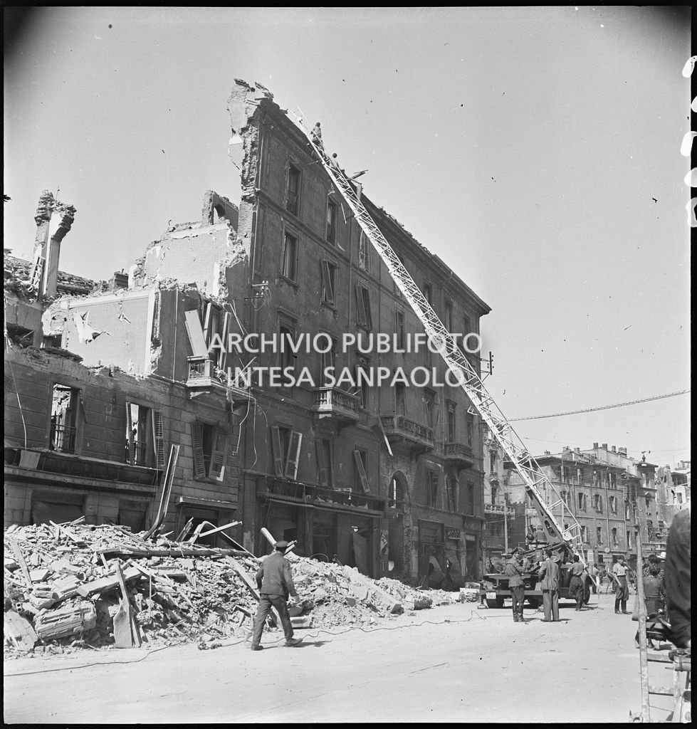 Due uomini verificano, con l'ausilio di un'autoscala, la stabilità di un edificio bombardato, in corso Genova a Milano, di cui ha resistito solo la facciata. A terra, addossati ai lati della strada, mucchi di macerie