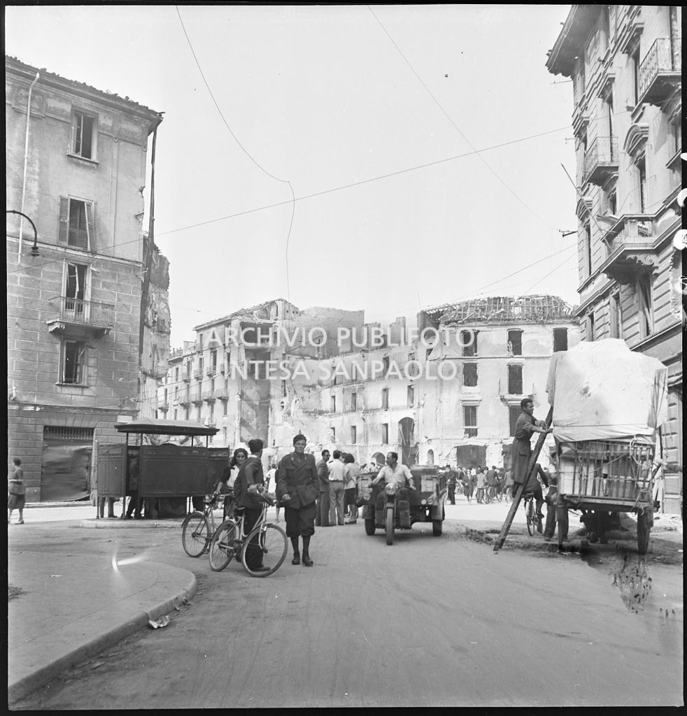 Passanti a piedi e in bicicletta, un motofurgone e un carro, in via Giuseppe Prina a Milano all'indomani dei bombardamenti che hanno gravemente danneggiato gli edifici