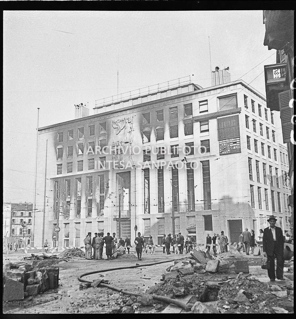 Palazzo del Popolo d'Italia, poi Palazzo dell'Informazione, in piazza Cavour a Milano, danneggiato dai bombardamenti. Sopra l'ingresso le scritte: "W il re W l'Italia", a destra, sempre in facciata, il datario segna il 28 luglio e sopra di esso la scritta: "Viva l'Italia". La piazza è ingombra di macerie e si notano capannelli di persone e militari