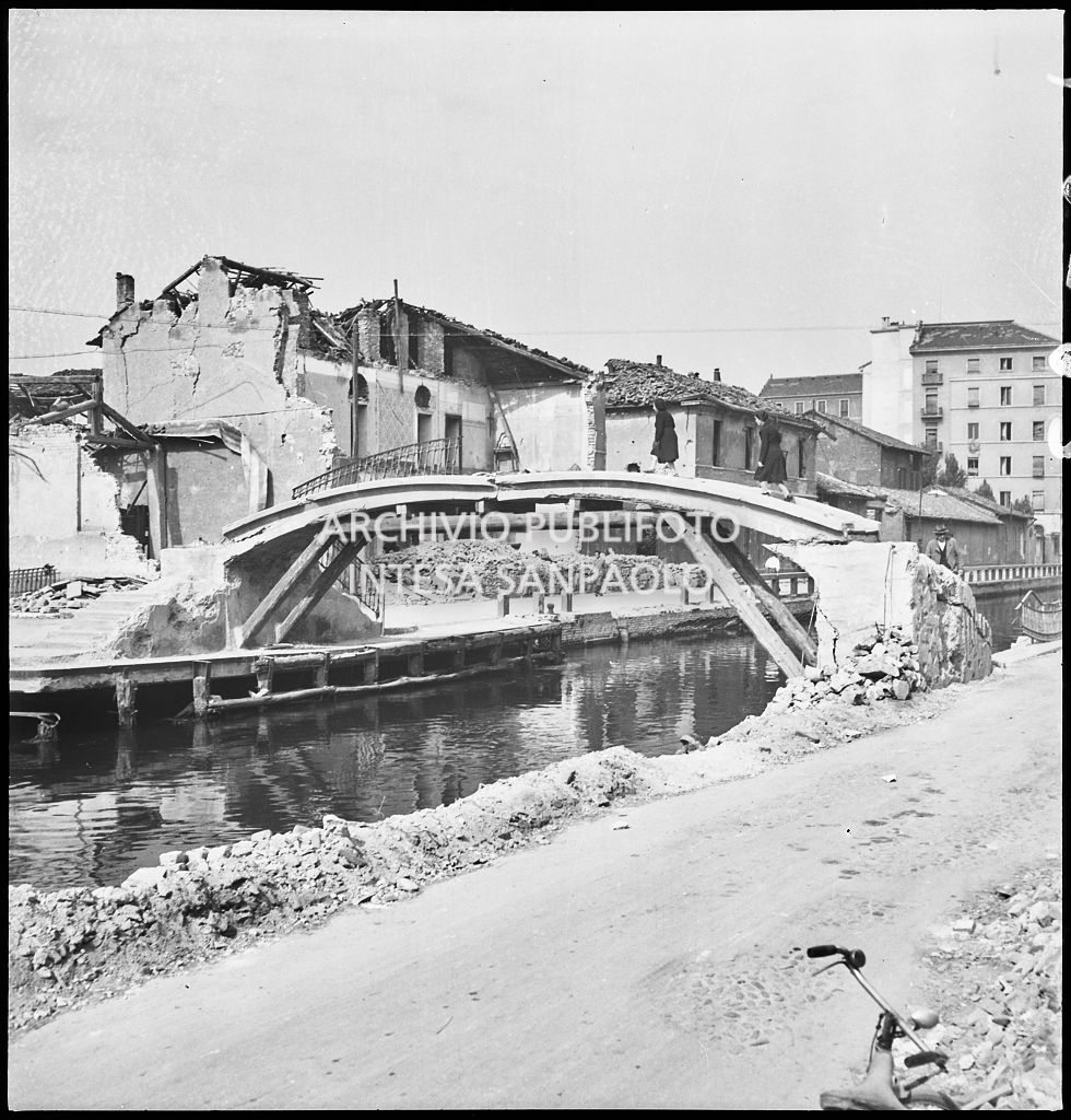 Edifici bombardati lungo il Naviglio Pavese; in primo piano due donne attraversano un ponte danneggiato ma che ha resistito agli attacchi aerei