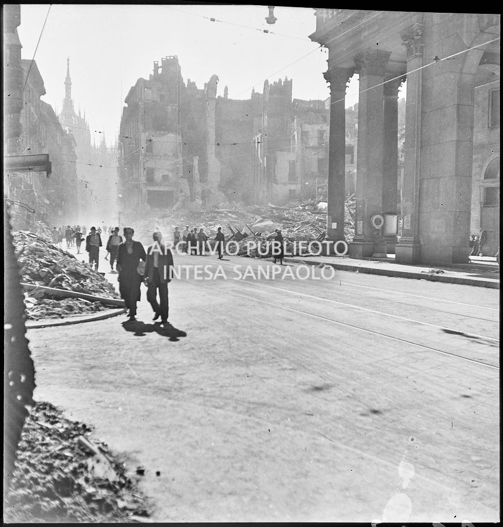 Scorcio di corso Vittorio Emanuele II a Milano all'indomani dei bombardamenti: passanti camminano per strada, sullo sfondo il Duomo, gli edifici distrutti durante gli attacchi aerei e piazza San Carlo ingombra di macerie