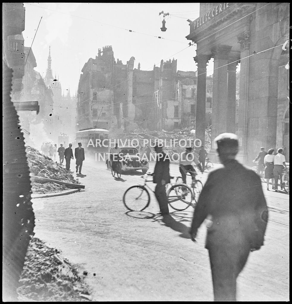 Scorcio di corso Vittorio Emanuele II a Milano all'indomani dei bombardamenti: tram tipo 1928, automobili e biciclette avanzano nella via, passanti camminano per strada, sullo sfondo edifici distrutti durante gli attacchi aerei e piazza San Carlo ingombra di macerie