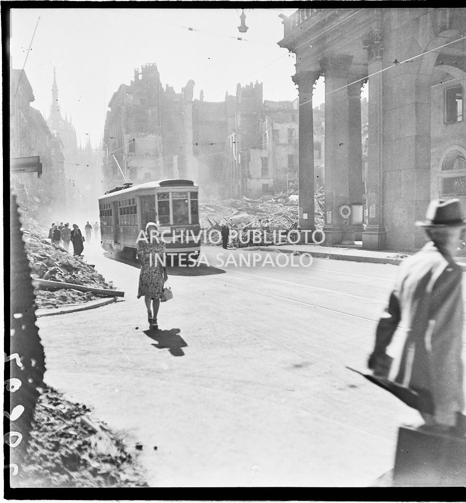 Scorcio di corso Vittorio Emanuele II a Milano all'indomani dei bombardamenti: un tram tipo 1928 avanza nella via, passanti camminano per strada, sullo sfondo edifici distrutti durante gli attacchi aerei e piazza San Carlo ingombra di macerie