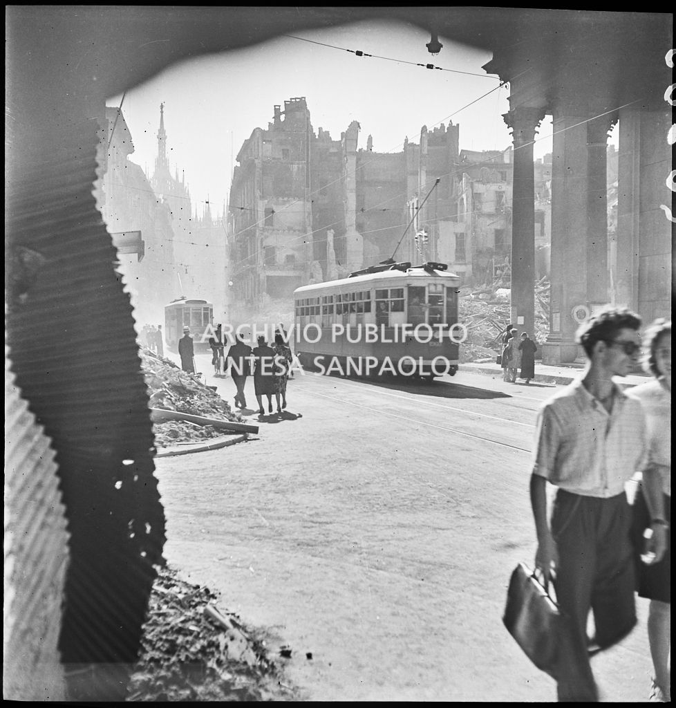 Scorcio di corso Vittorio Emanuele II a Milano all'indomani dei bombardamenti: tram tipo 1928 avanzano nella via, passanti camminano per strada, sullo sfondo edifici distrutti durante gli attacchi aerei e piazza San Carlo ingombra di macerie