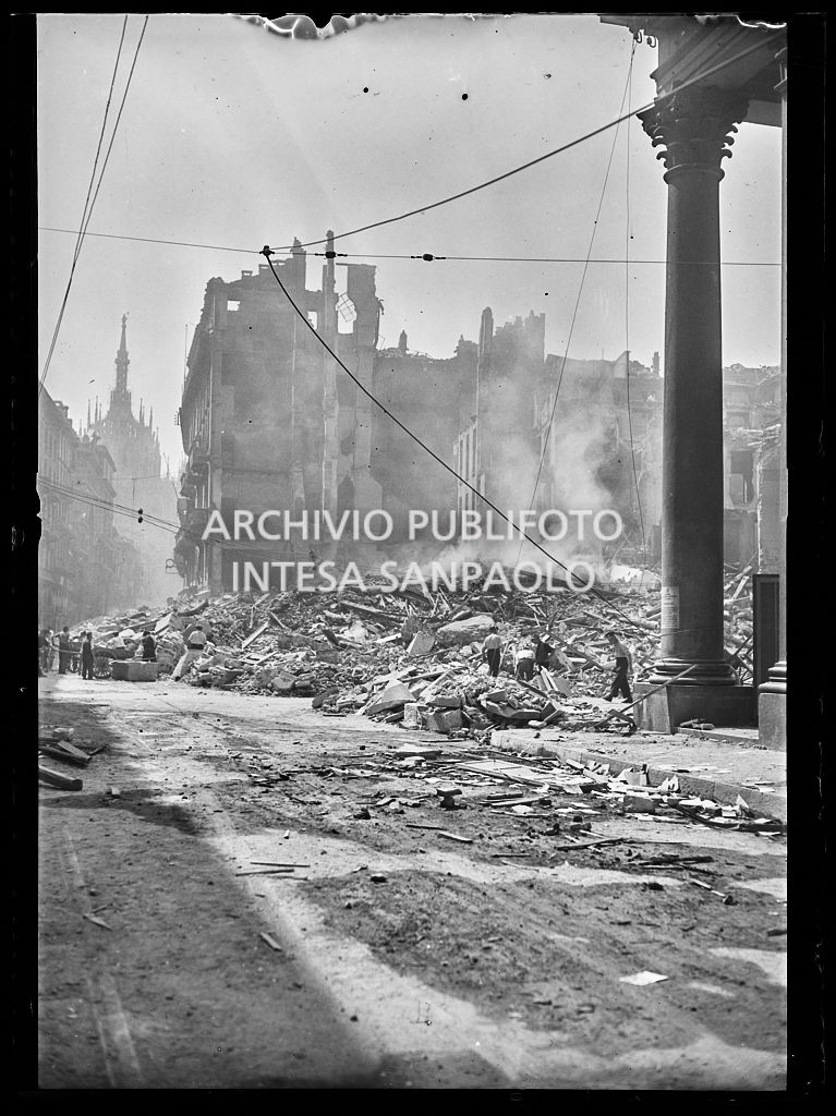 Scorcio di piazza San Carlo a Milano, all'indomani dei bombardamenti, ingombra delle macerie, ancora fumanti, degli edifici e della basilica di San Carlo al Corso, gravemente danneggiati durante gli attacchi aerei. Sullo sfondo il Duomo