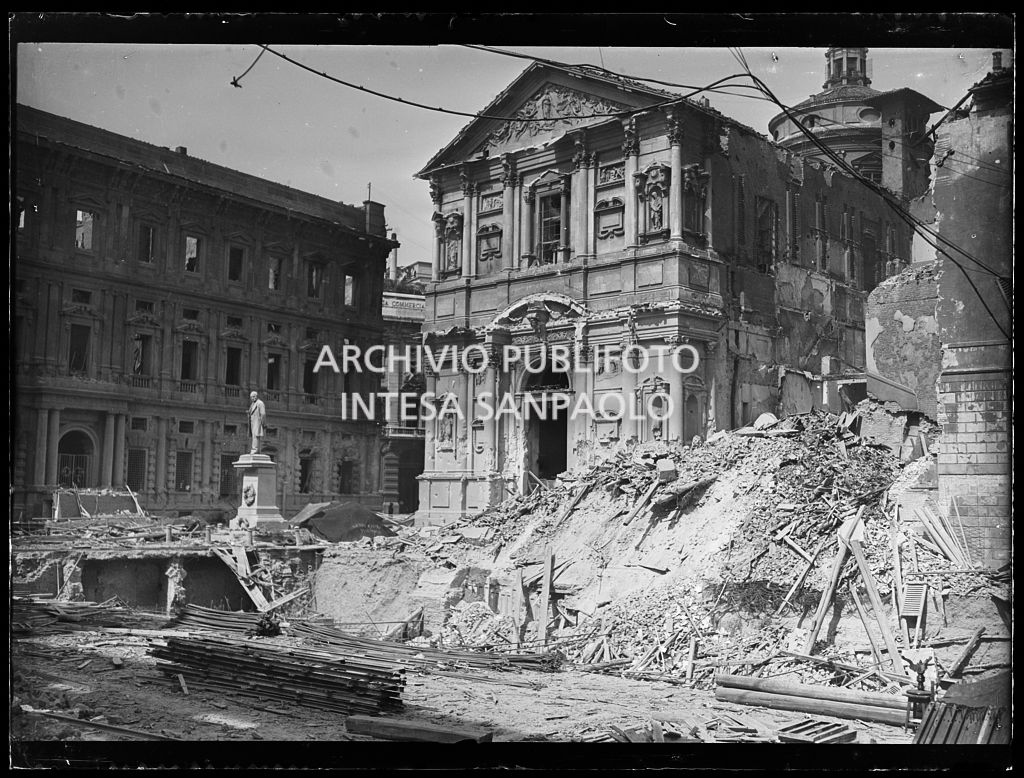 Facciata posteriore di Palazzo Marino, su piazza San Fedele a Milano, e la chiesa di San Fedele, entrambi gravemente danneggiati dai bombardamenti. A destra della chiesa il cumulo di macerie in cui fu ridotto l'edificio che ospitava la questura di Milano; in mezzo alla piazza il monumento a Alessandro Manzoni intatto.
