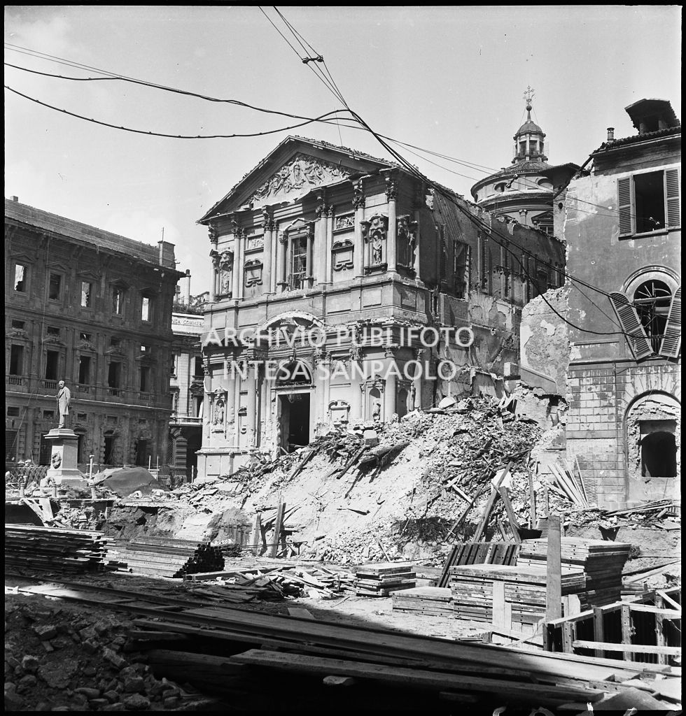 Facciata posteriore di Palazzo Marino, su piazza San Fedele a Milano, e la chiesa di San Fedele, entrambi gravemente danneggiati dai bombardamenti. A destra della chiesa il cumulo di macerie in cui fu ridotto l'edificio che ospitava la questura di Milano; in mezzo alla piazza il monumento a Alessandro Manzoni intatto.