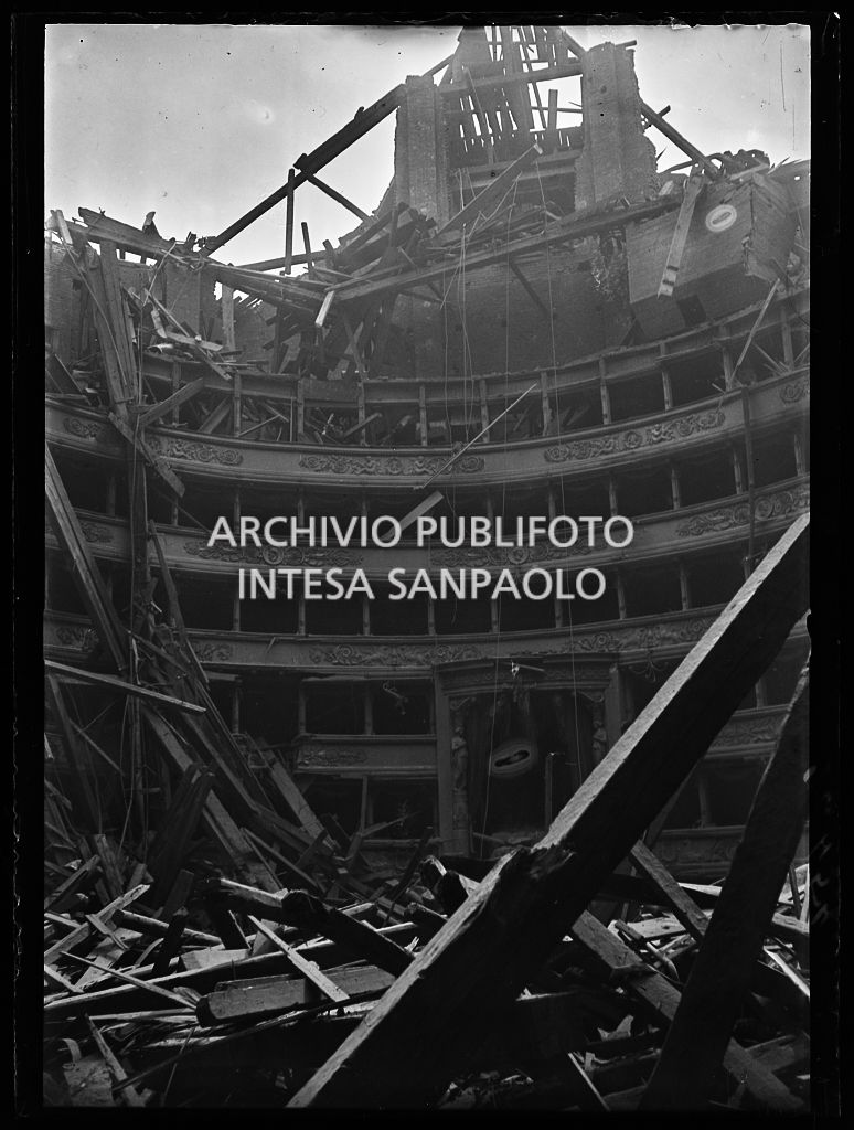 Scorcio dell'interno del Teatro alla Scala di Milano dopo i bombardamenti: il tetto e la volta centinata sottostante sono completamente crollati distruggendo in parte il quinto e sesto ordine di gallerie e danneggiando i palchi, il proscenio e la platea. Sullo sfondo il palco reale