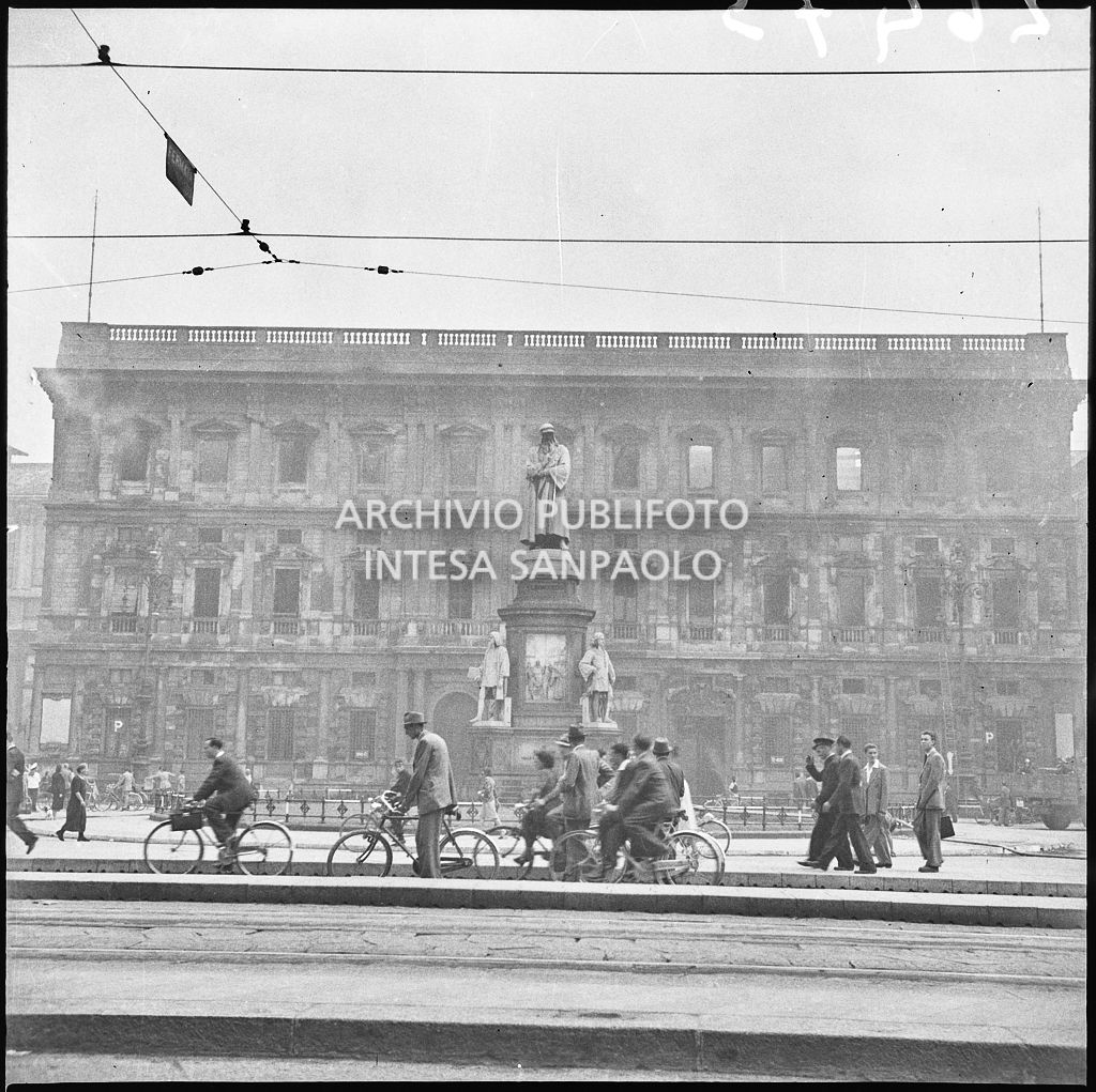 Passanti a piedi e in bicicletta in piazza della Scala a Milano, al cui centro si erge, illeso, il monumento a Leonardo da Vinci. Sullo sfondo Palazzo Marino, a Milano, completamente bruciato e ancora fumante a causa dei bombardamenti, di cui sono rimasti intatti solo i muri perimetrali. Sulla destra, appoggiata al palazzo, la scala dei vigili del fuoco