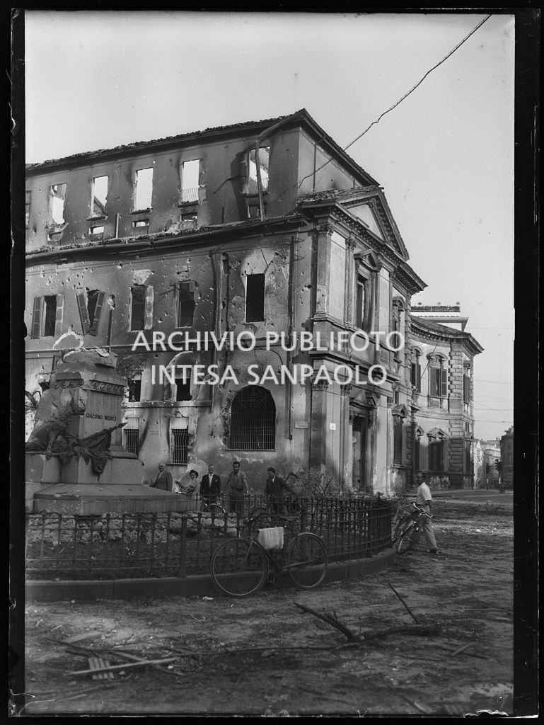 Alcune persone, in via Senato a Milano, intorno alla recinzione che proteggeva il monumento a Giacomo Medici distrutto dai bombardamenti che hanno gravemente danneggiato anche Palazzo del Senato alle loro spalle