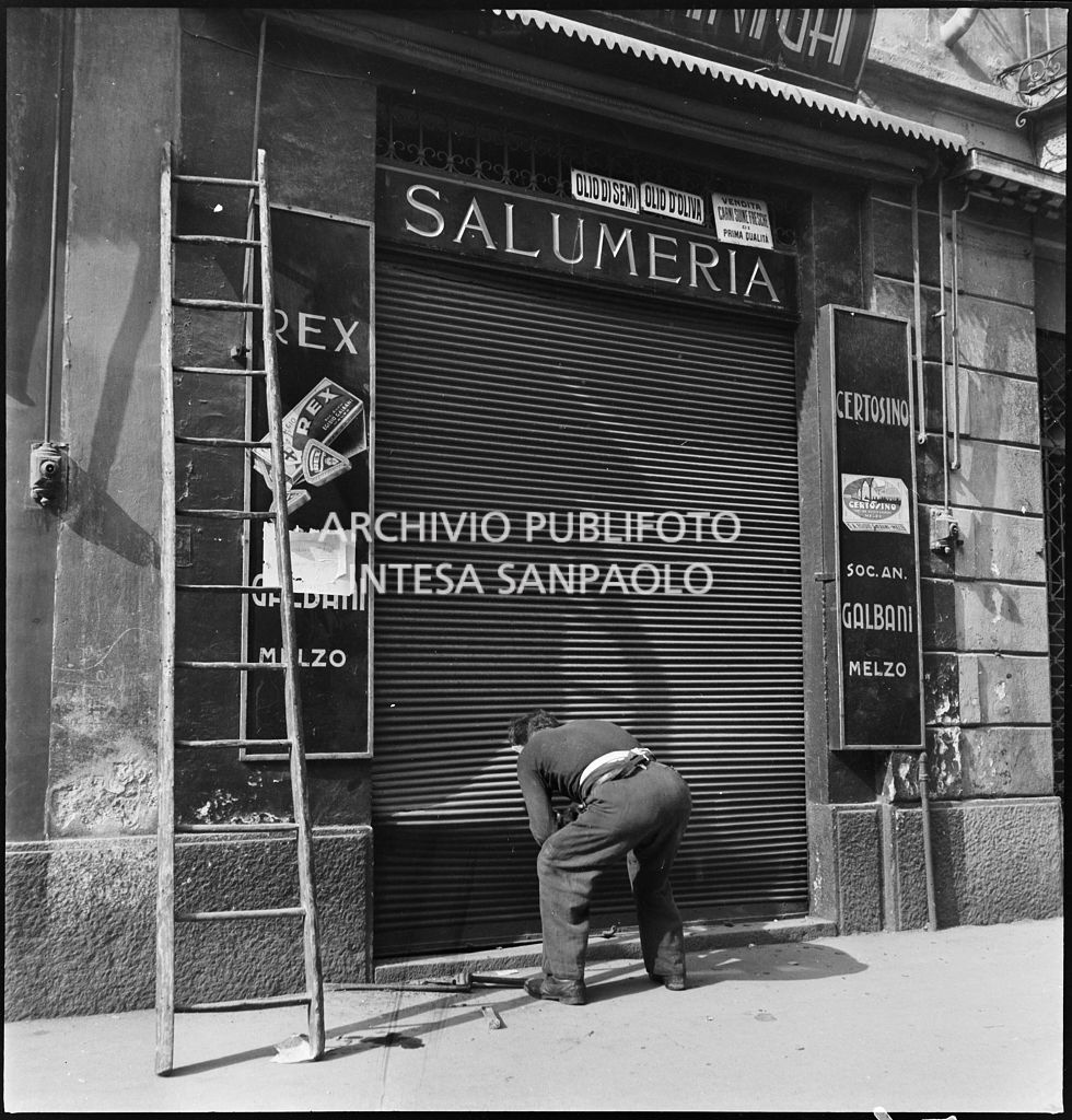Un uomo piegato sulla saracinesca , deformata dai bombardamenti, di una salumeria deformate dai bombardamenti