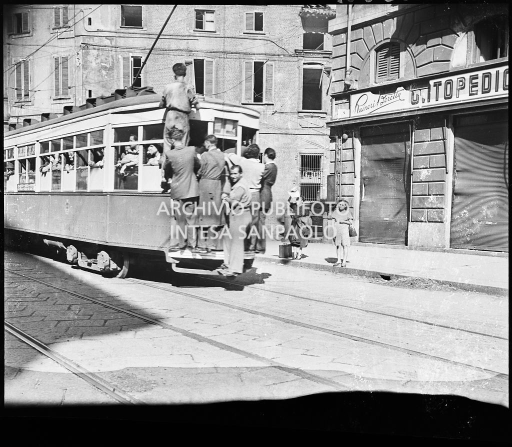 Tram tipo 1928 in transito in corso Venezia a Milano nei giorni dei bombardamenti della città; la vettura è così affollata che alcuni passeggeri viaggiano in piedi sul paraurti posteriore (dipinto di bianco per aumentarne la visibilità durante l'oscuramento), sul gancio e uno si è addirittura arrampicato sul finestrino. Sulla destra il negozio di articoli ortopedici Raineri Beretta