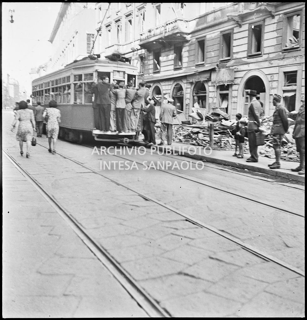 Tram tipo 1928 in transito in corso Venezia a Milano nei giorni dei bombardamenti della città; la vettura è così affollata che alcuni passeggeri viaggiano in piedi sul paraurti posteriore (dipinto di bianco per aumentarne la visibilità durante l'oscuramento). Sul marciapiede le macerie degli edifici danneggiati durante gli attacchi aerei