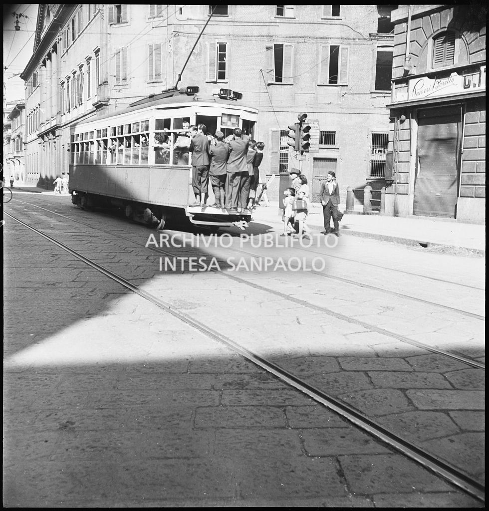 Tram tipo 1928 in transito in corso Venezia a Milano nei giorni dei bombardamenti della città; la vettura è così affollata che alcuni passeggeri viaggiano in piedi sul paraurti posteriore (dipinto di bianco per aumentarne la visibilità durante l'oscuramento). Sulla destra il negozio di articoli ortopedici Raineri Beretta, sullo sfondo Palazzo Serbelloni
