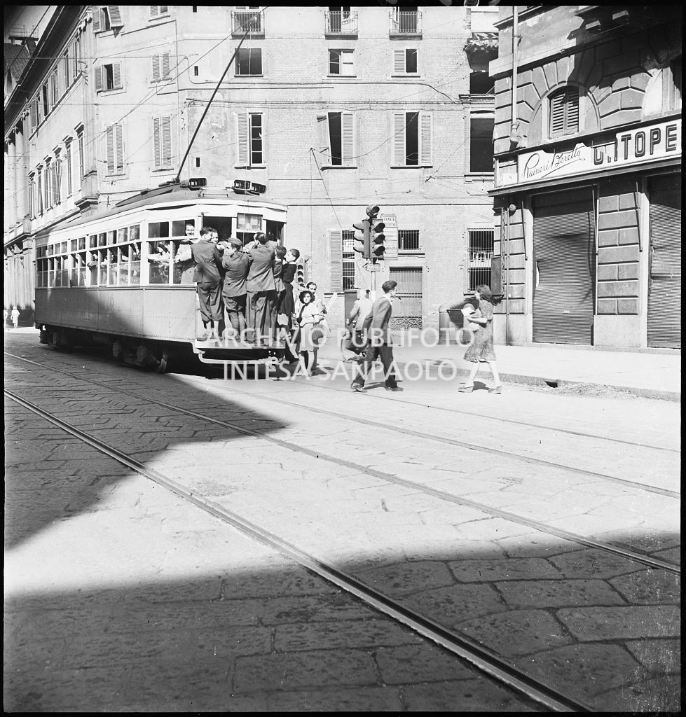 Tram tipo 1928 in transito in corso Venezia a Milano nei giorni dei bombardamenti della città; la vettura è così affollata che alcuni passeggeri viaggiano in piedi sul paraurti posteriore (dipinto di bianco per aumentarne la visibilità durante l'oscuramento). Sulla destra il negozio di articoli ortopedici Raineri Beretta