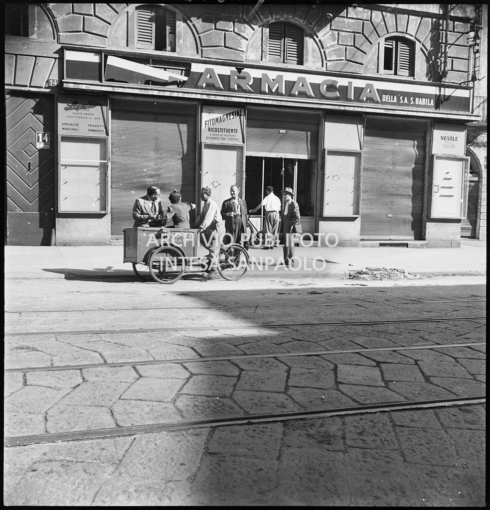 Un uomo alla guida di una bicicletta con cassone per trasporto medicinali, della Sandoz, all'esterno della Farmacia della Società Anonima S. Babila, in corso Venezia a Milano, danneggiata durante i bombardamenti