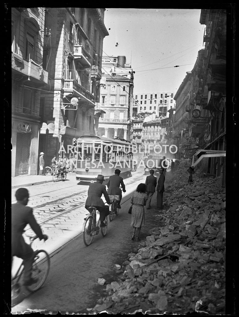 Scorcio di corso Vittorio Emanuele II, a Milano, all'indomani dei bombardamenti: le macerie degli edifici distrutti durante gli attacchi aerei sono state addossate ai lati della strada per consentire la circolazione a tram, automobili biciclette, autocarri e pedoni come in questa immagine. Un tram tipo 1928, linea 22, avanza nella via